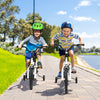 Two kids riding 14-inch white bikes with training wheels, enjoying a sunny day by the park near a lake.