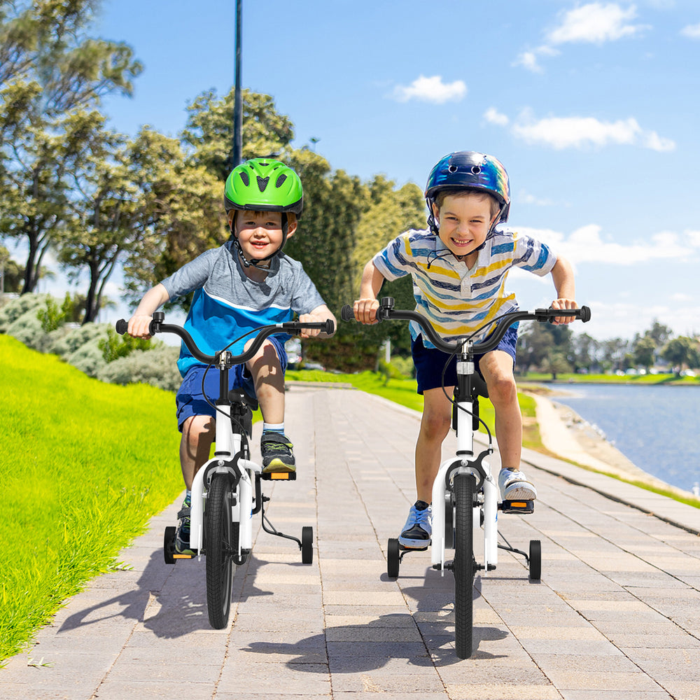 Two kids riding 14-inch white bikes with training wheels, enjoying a sunny day by the park near a lake.