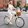 Side view of a woman cycling on a 24-inch beige beach cruiser bike with a stylish woven basket and cup holder.