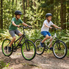 Two kids riding 24-inch adjustable mountain bikes with 7-speed gears and suspension forks on a forest trail.