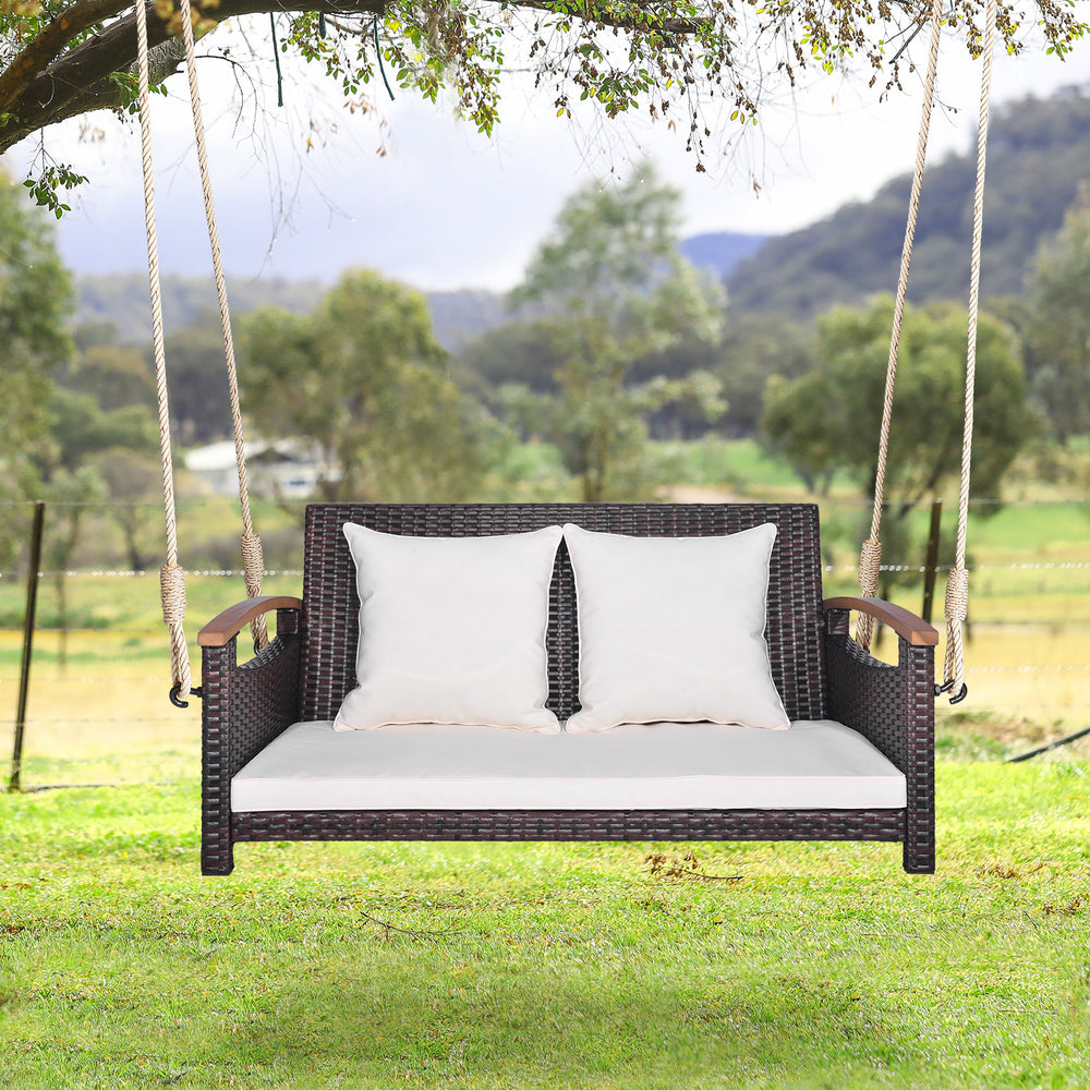 2-person patio rattan porch swing with cushions, hanging from a tree, perfect for backyard relaxation.