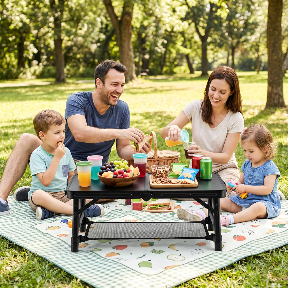 Folding camping table in black, set up for a picnic with family, featuring drinks and snacks on a grassy area.
