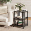 3-tier rolling end table in black with open storage shelves, placed beside a cozy sofa and decorated with flowers.