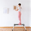 A woman standing on a folding aluminum step stool ladder, using it to hang a picture frame on the wall.
