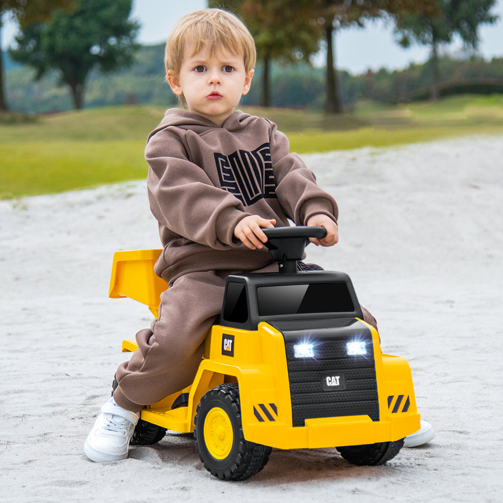 Child riding a yellow 6V electric Caterpillar construction vehicle with LED lights and detachable dump bed on a sandy.