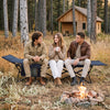 Camping cot for adults with soft padded mat, navy color, shown in a cozy outdoor setting by a campfire.