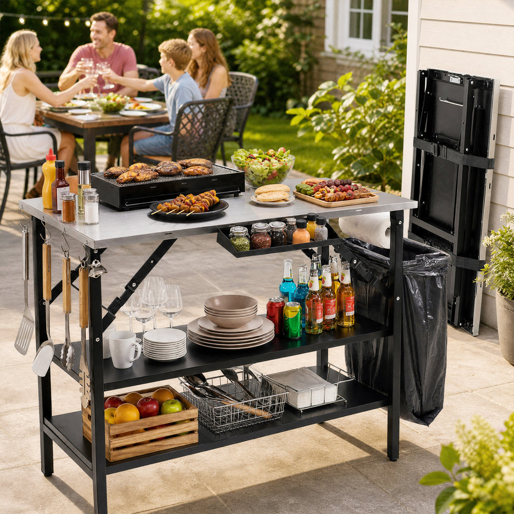 Foldable outdoor cook station in black with stainless steel top, removable shelves, and trash bag holder for convenient cooking.