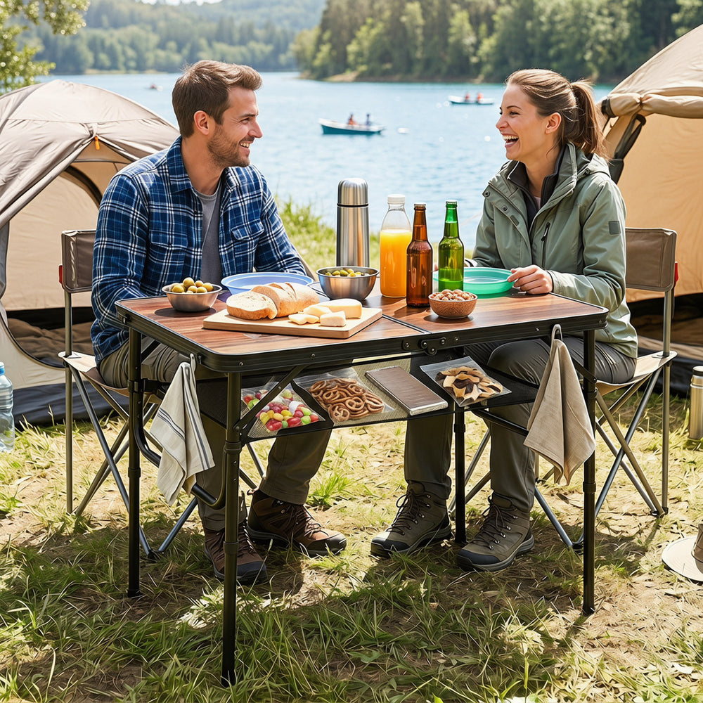 Folding camping table in black and brown, set up by a lake with drinks and snacks, featuring adjustable heights and.