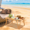 Folding camping table with waterproof surface, brown finish, set up for a picnic on the beach with food and drinks.