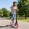 Girl riding a pink kick scooter with large light-up wheels in a park, suitable for kids and teens aged 8+.