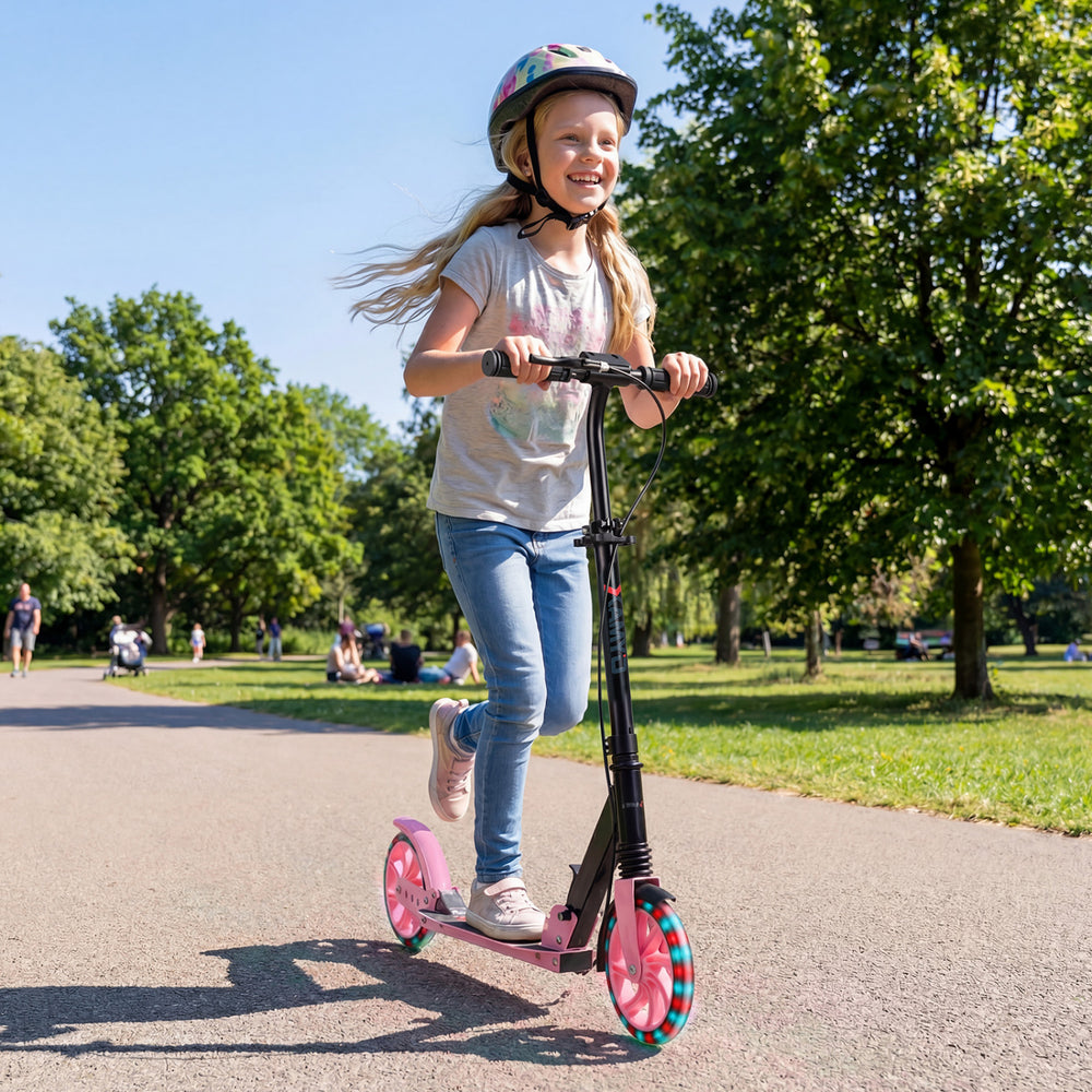 Girl riding a pink kick scooter with large light-up wheels in a park, suitable for kids and teens aged 8+.