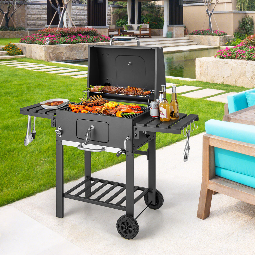 Outdoor BBQ charcoal grill with foldable side tables, wheels, and a large cooking area for backyard gatherings.