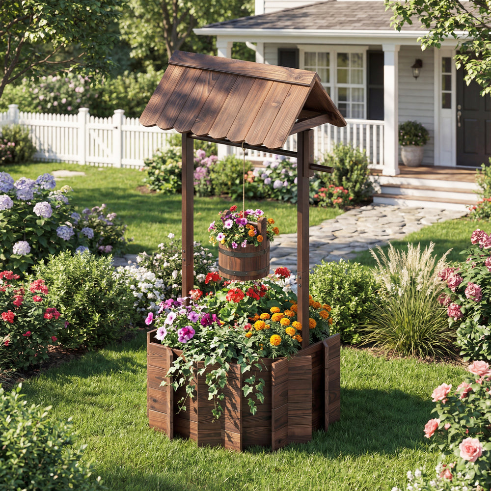 Outdoor wooden wishing well planter with rustic burnt-like finish, featuring a bucket for smaller plants in a garden.