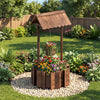 Outdoor wooden wishing well planter with burnt-like finish, featuring a hanging bucket for flowers in a garden setting.