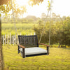 Single rattan porch swing with white cushion, featuring acacia wood armrests, hanging in a sunny outdoor setting.