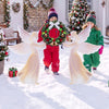 Two children in winter attire carry a festive wreath alongside pink PVC angel decorations in a snowy outdoor setting.