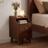 Wood nightstand with two drawers and solid wood legs in brown, styled beside a bed with decorative lamp and books.