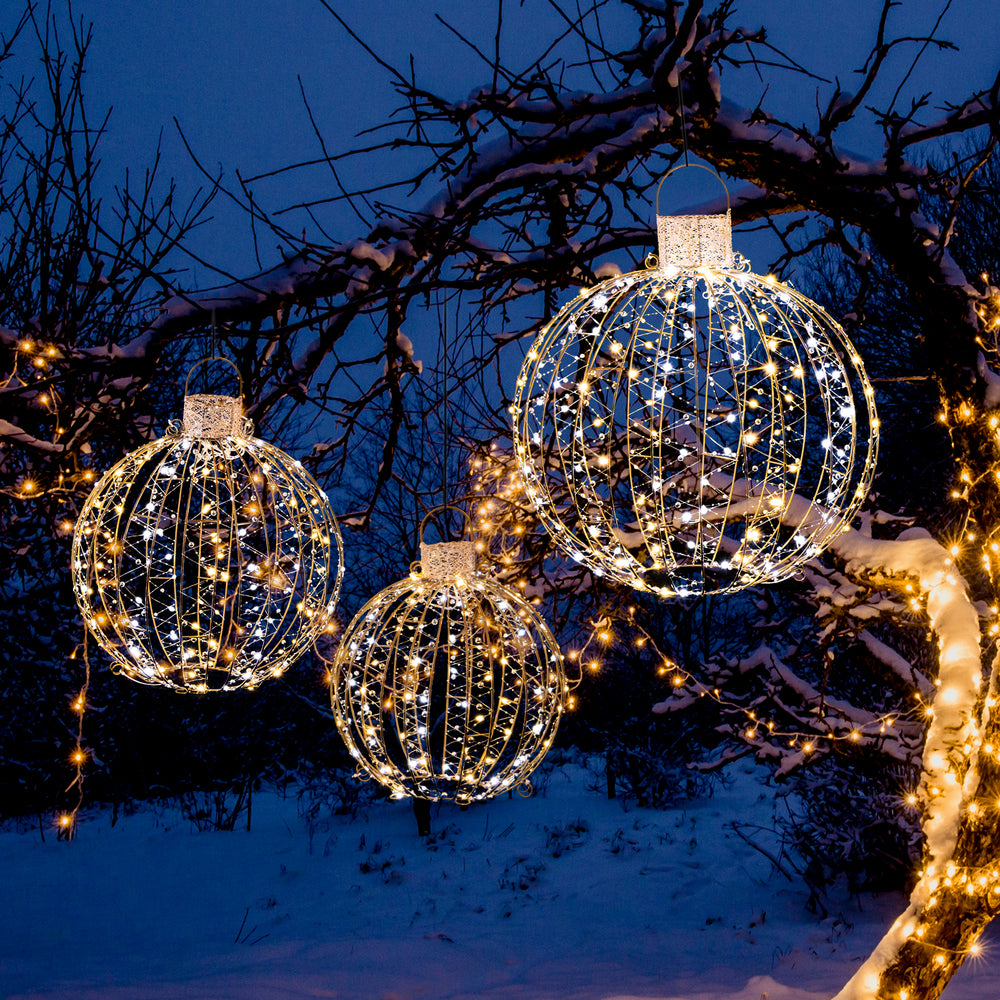 Three decorative Christmas LED light balls in different sizes, featuring warm and white lights, hung on a snowy tree.