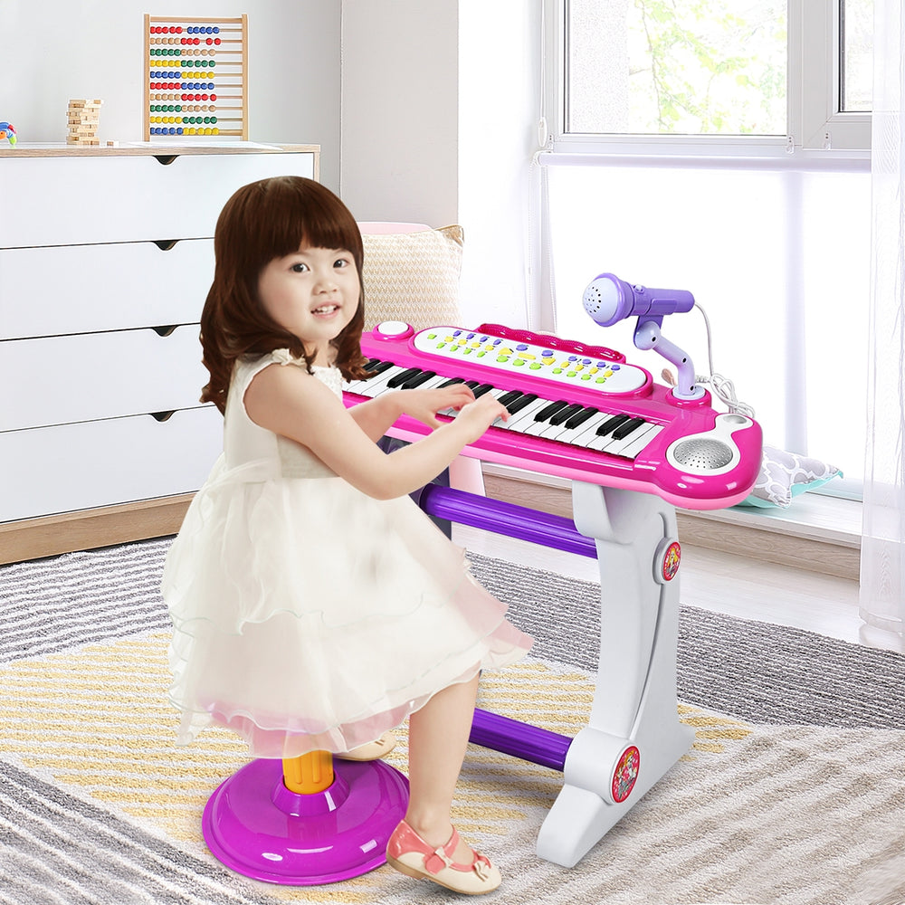 Child playing the pink 37 key electronic keyboard piano with detachable microphone and stool in a bright room.