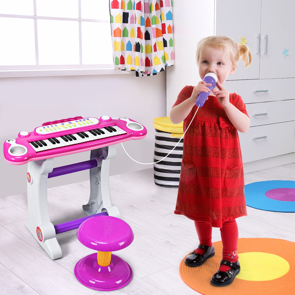 Child playing with a pink 37 key electronic keyboard toy piano, featuring a detachable microphone and stool for fun.