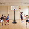 Adjustable portable basketball hoop with shatterproof backboard, shown in use by players in a gym setting.